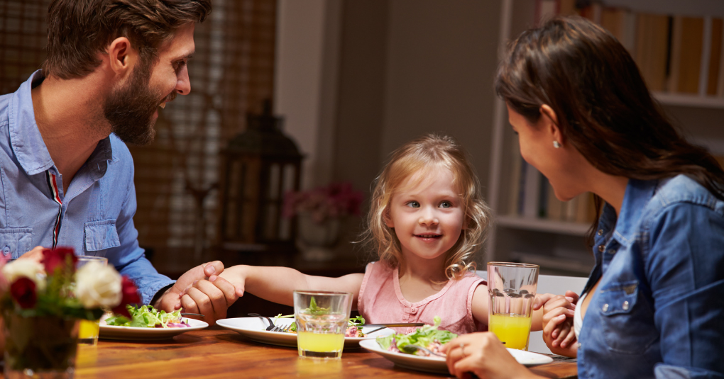 Family at dining table with food surrounding while holding hands.
